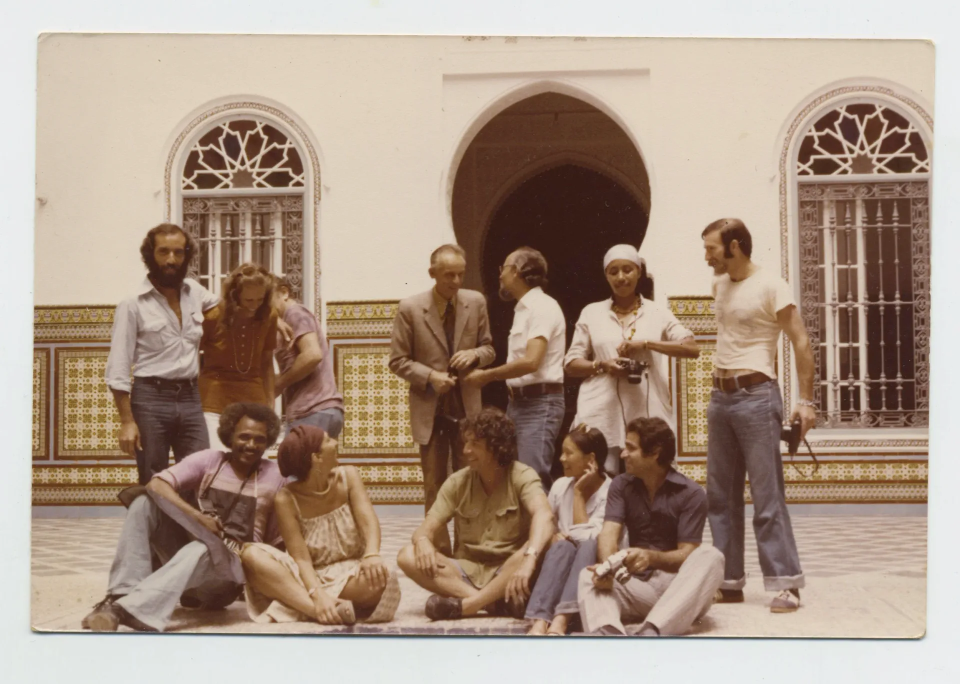 Color photograph of a group of people posed and seated in front of a colonial-style building with ornate arched doorways and tiled facade.