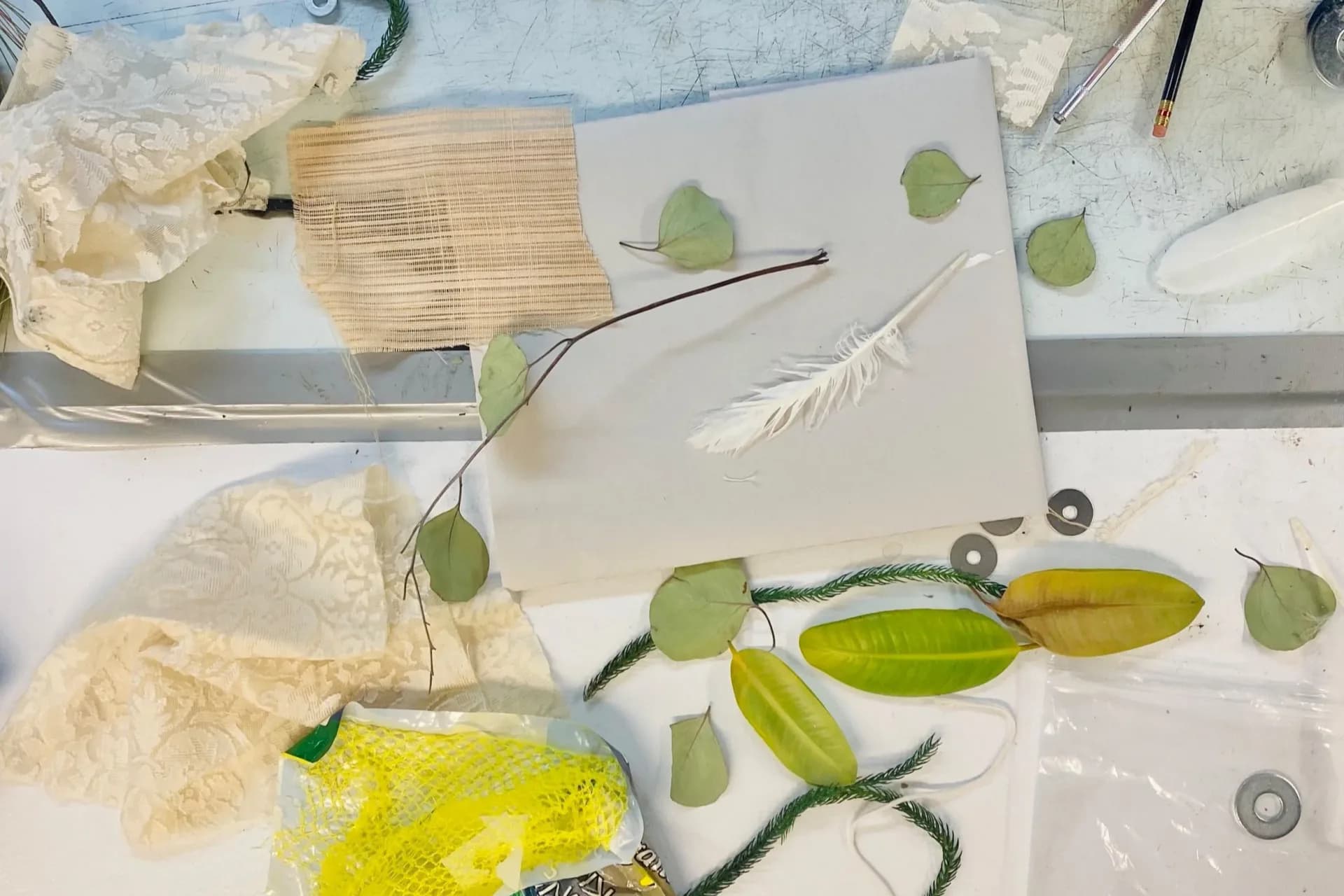 Mixed-media artwork featuring pressed green leaves, flowers, and plant materials arranged on white paper with fabric and string elements.
