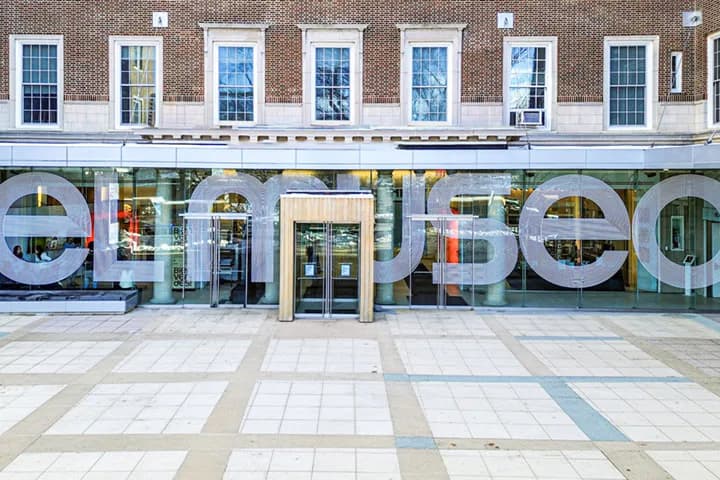 Storefront exterior of El Museo del Barrio with large circular signage, glass windows, brick façade above, and geometric paving pattern below.