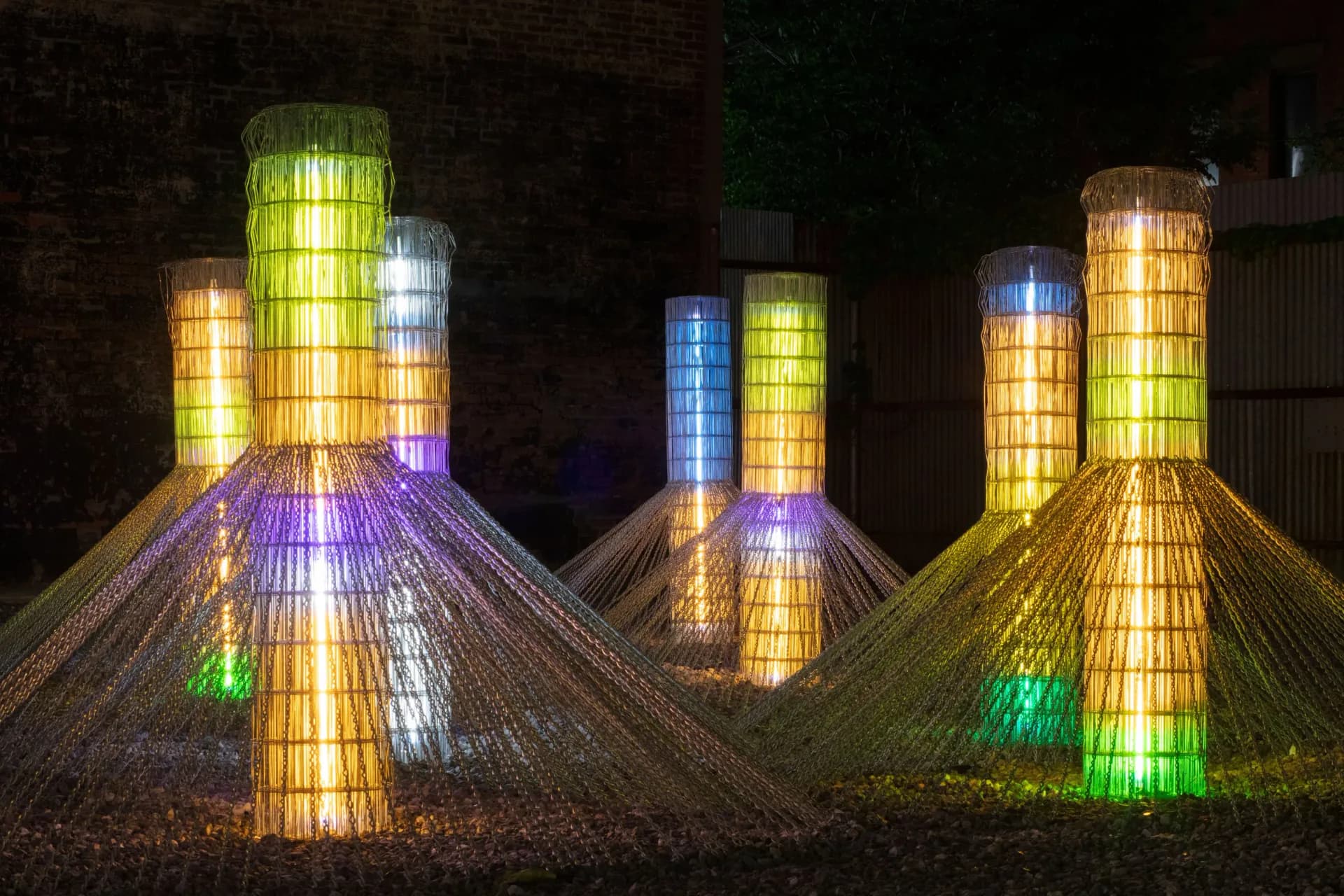 Illuminated outdoor light installation with stacked cylindrical mesh towers in yellow, green, blue, and orange, creating radiating light patterns on the ground.