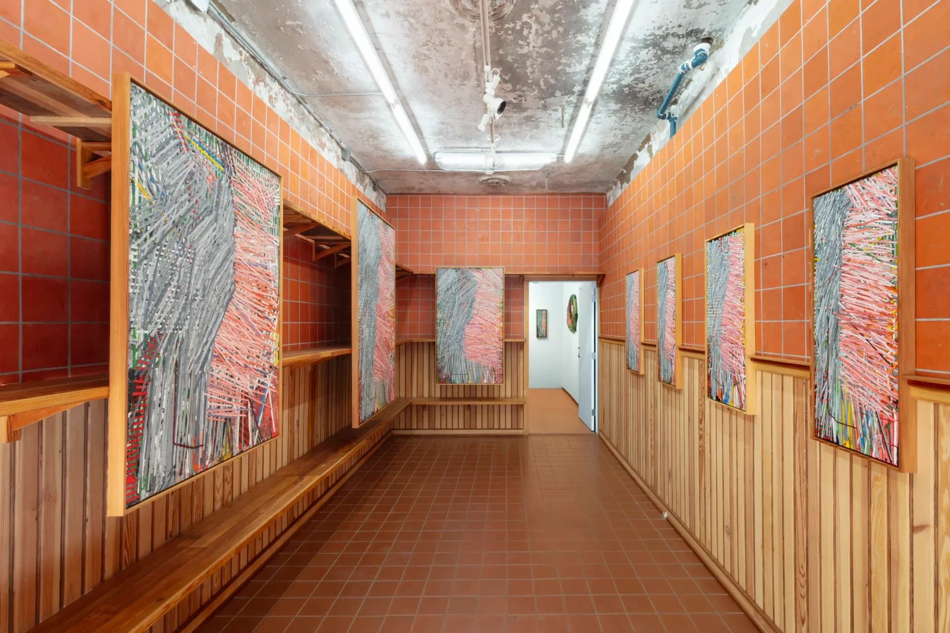 Gallery corridor with orange tile walls, wooden baseboards, framed artworks displayed on both sides, and fluorescent ceiling lighting.