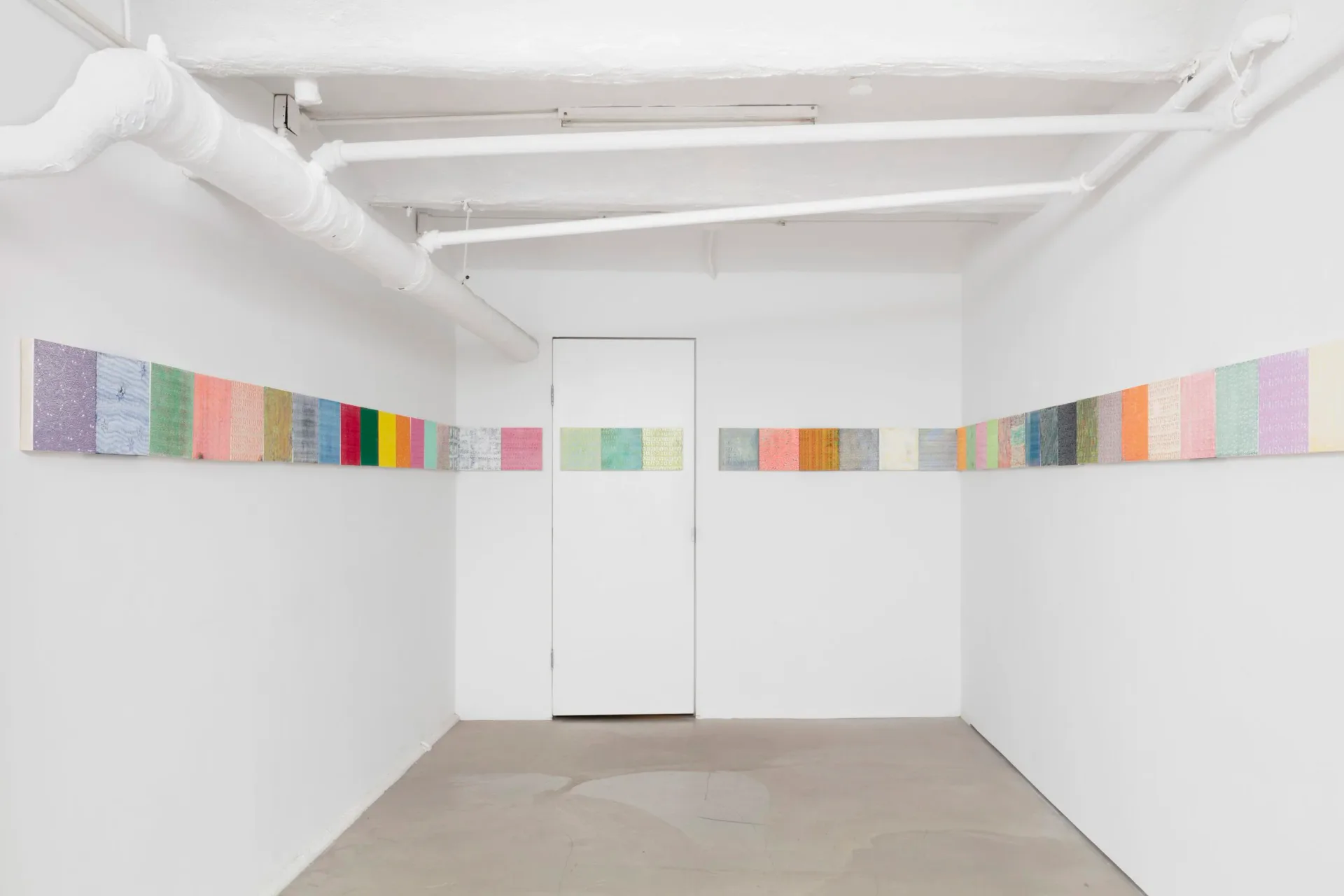 Minimalist white gallery space with concrete floor and a frieze of small colorful textile or paper squares running horizontally along both walls.