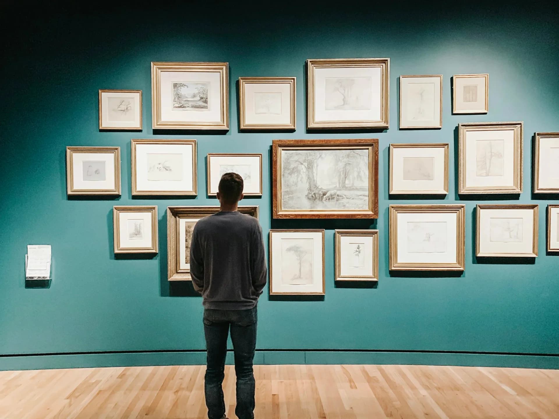 Person viewing gallery wall with arrangement of framed artwork and sketches on teal-colored wall in museum or gallery interior.