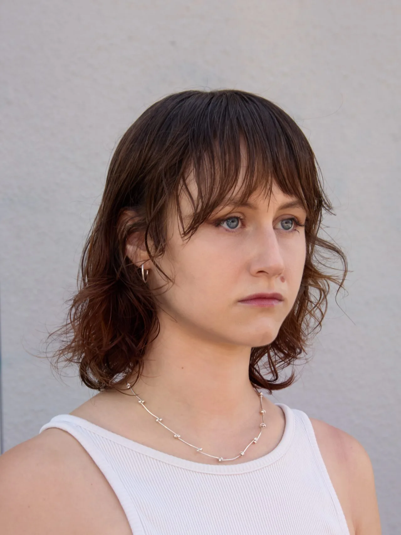 Young woman with shoulder-length wavy brown hair and bangs, wearing a white tank top and thin necklace, looking to the side against a plain gray backdrop