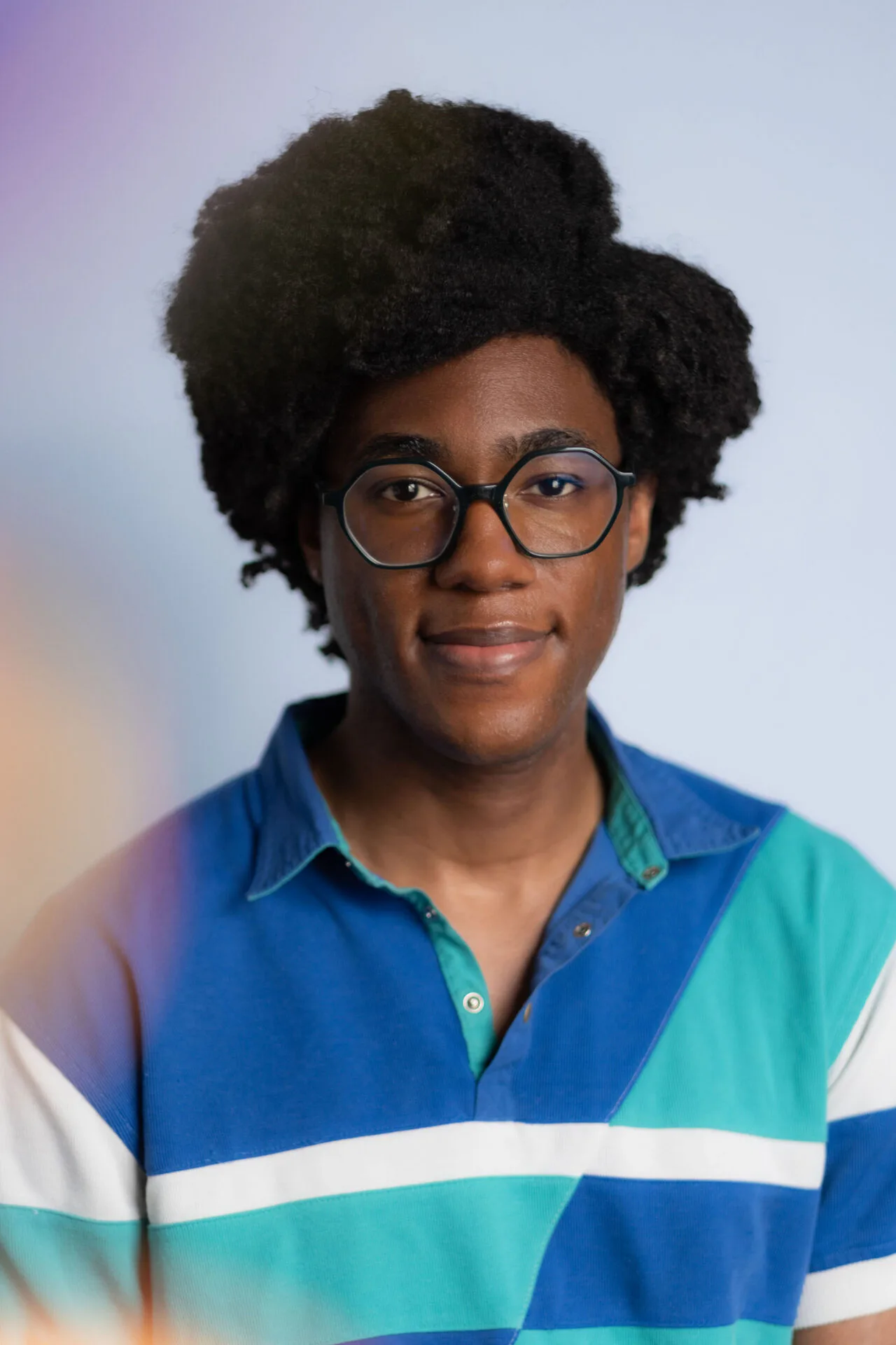 Young man with a curly afro and round glasses wearing a blue, teal and white striped rugby shirt, smiling gently against a soft blurred background