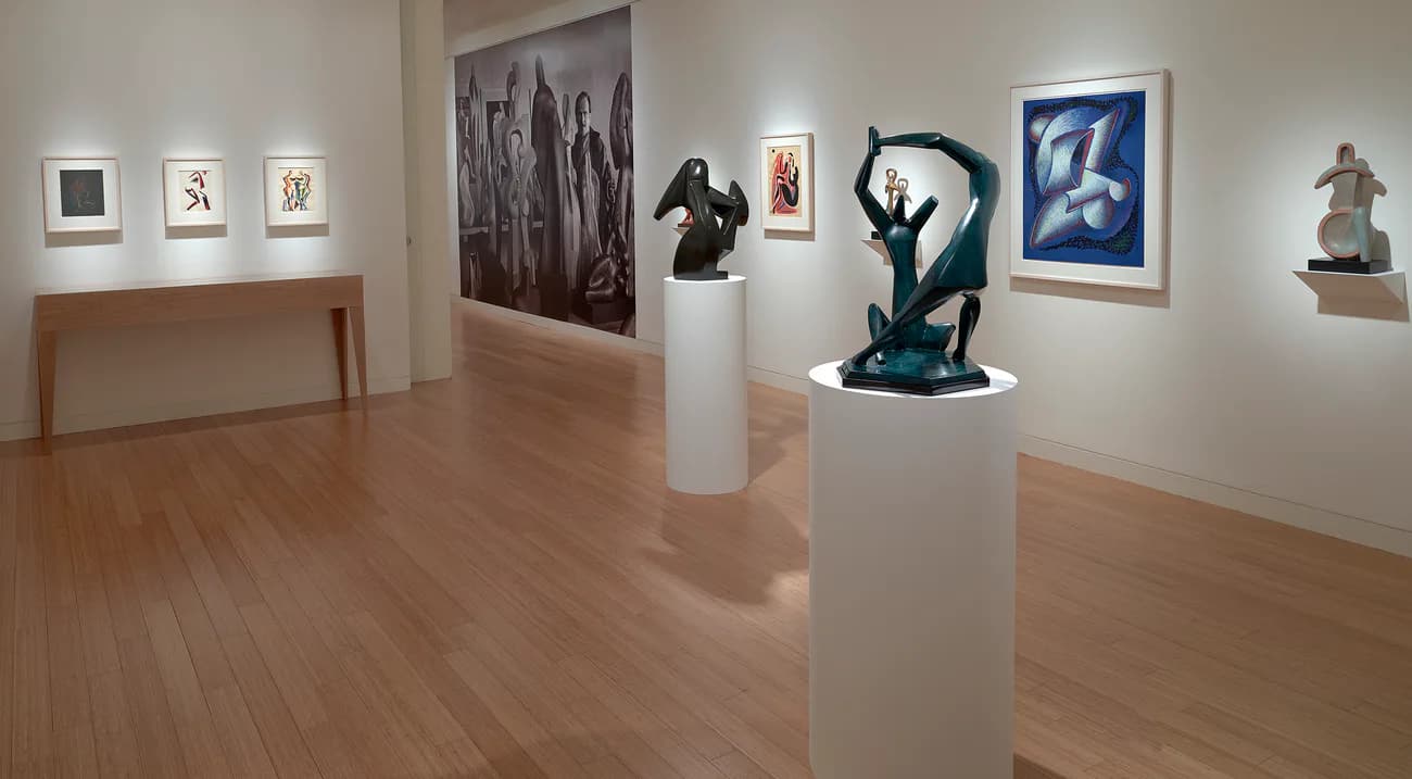 Gallery space with wooden floors featuring sculptures on pedestals, framed photographs, and wall-mounted artworks in mixed media.
