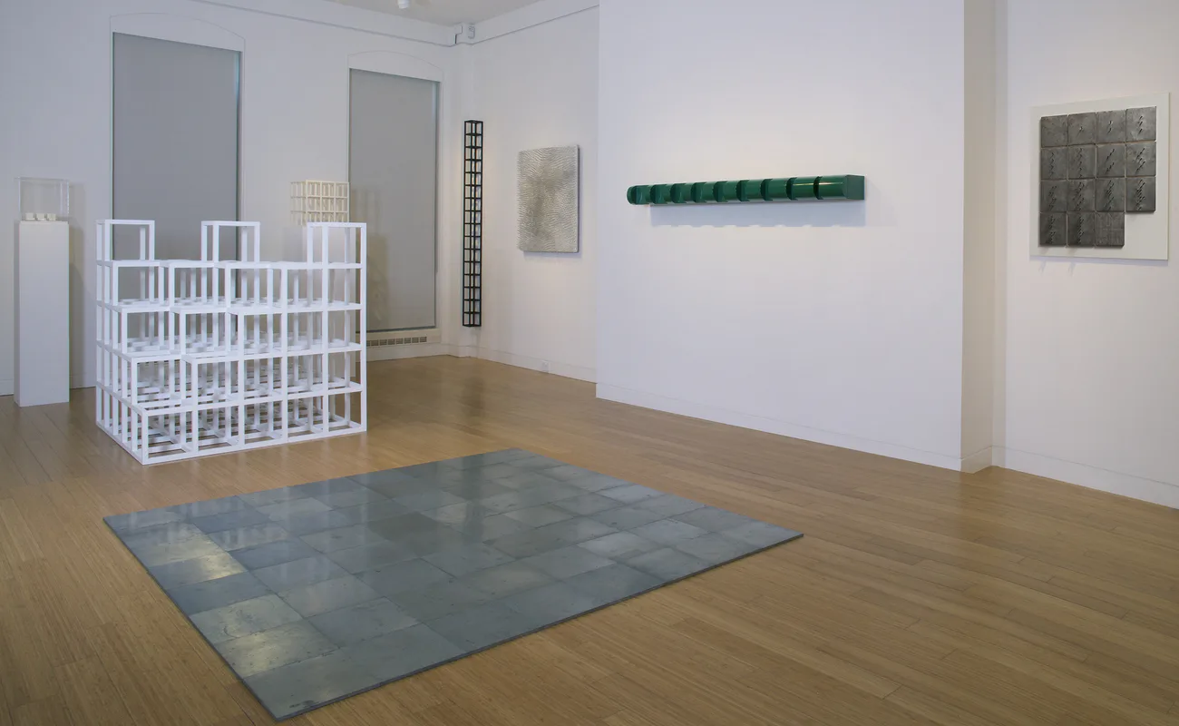 Minimalist gallery with white walls, wooden floors, white sculptural structure, blue-gray floor mat, and hanging green textile piece.