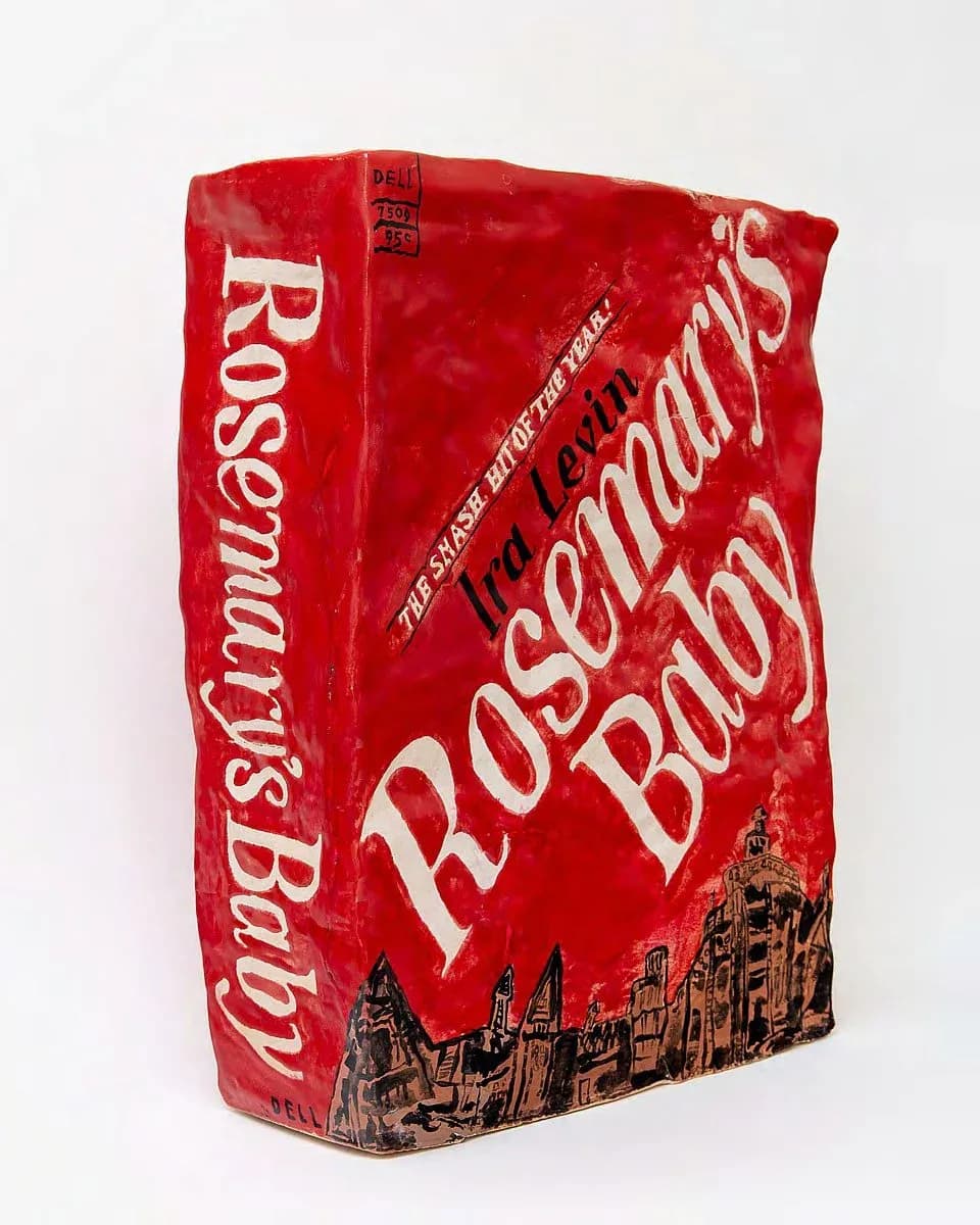 Sculptural object: bright red crumpled package or wrapper with white text reading "Rosemary's Baby" and architectural building imagery, three-dimensional form.