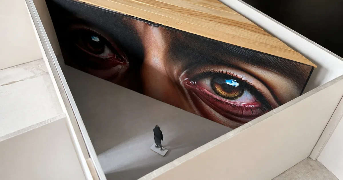 Gallery interior with white walls and wooden ceiling; large framed artwork of a close-up eye visible above, small figure visible on white floor below.