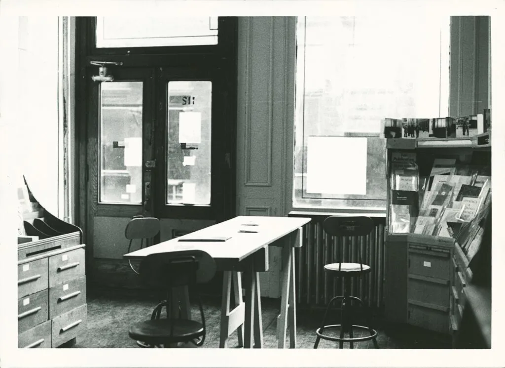 Interior office space with drafting table, stools, glass-paneled doors, tall windows, and a rack of publications