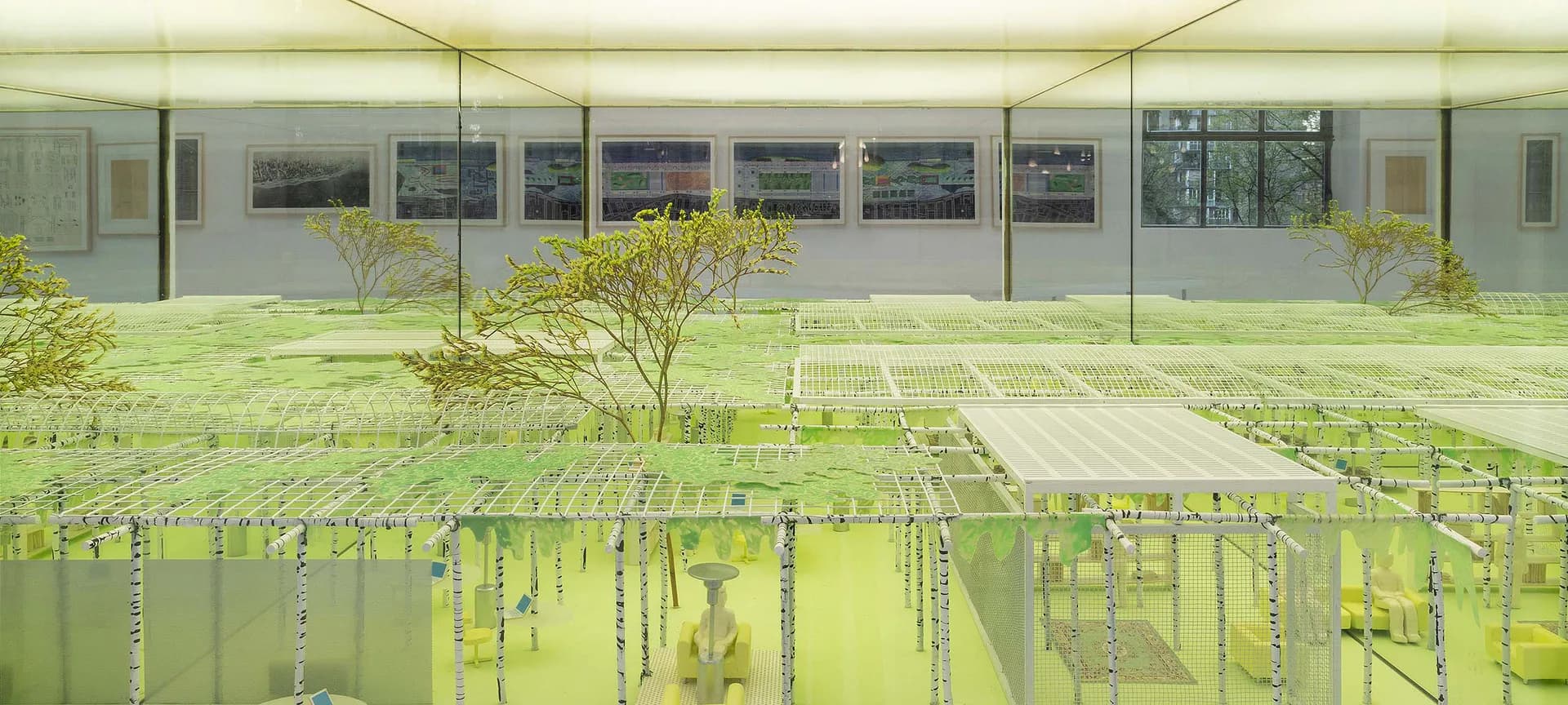 Interior gallery space with glass facade, featuring living plant installation with hanging green vegetation and suspended wires.
