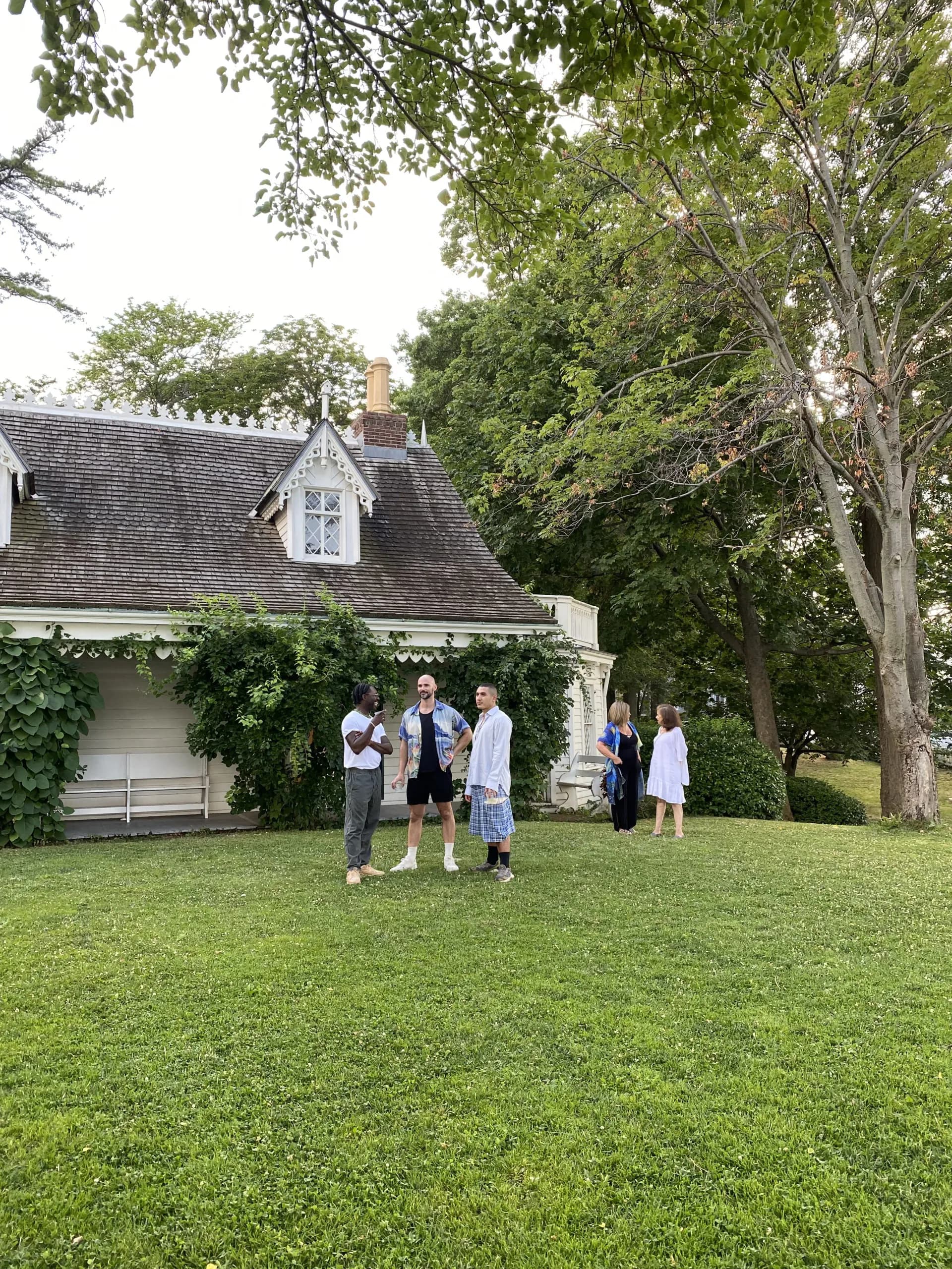 Exterior of the Alice Austen House cottage with gabled roof and dormer window, surrounded by trees and a green lawn where visitors are gathered.