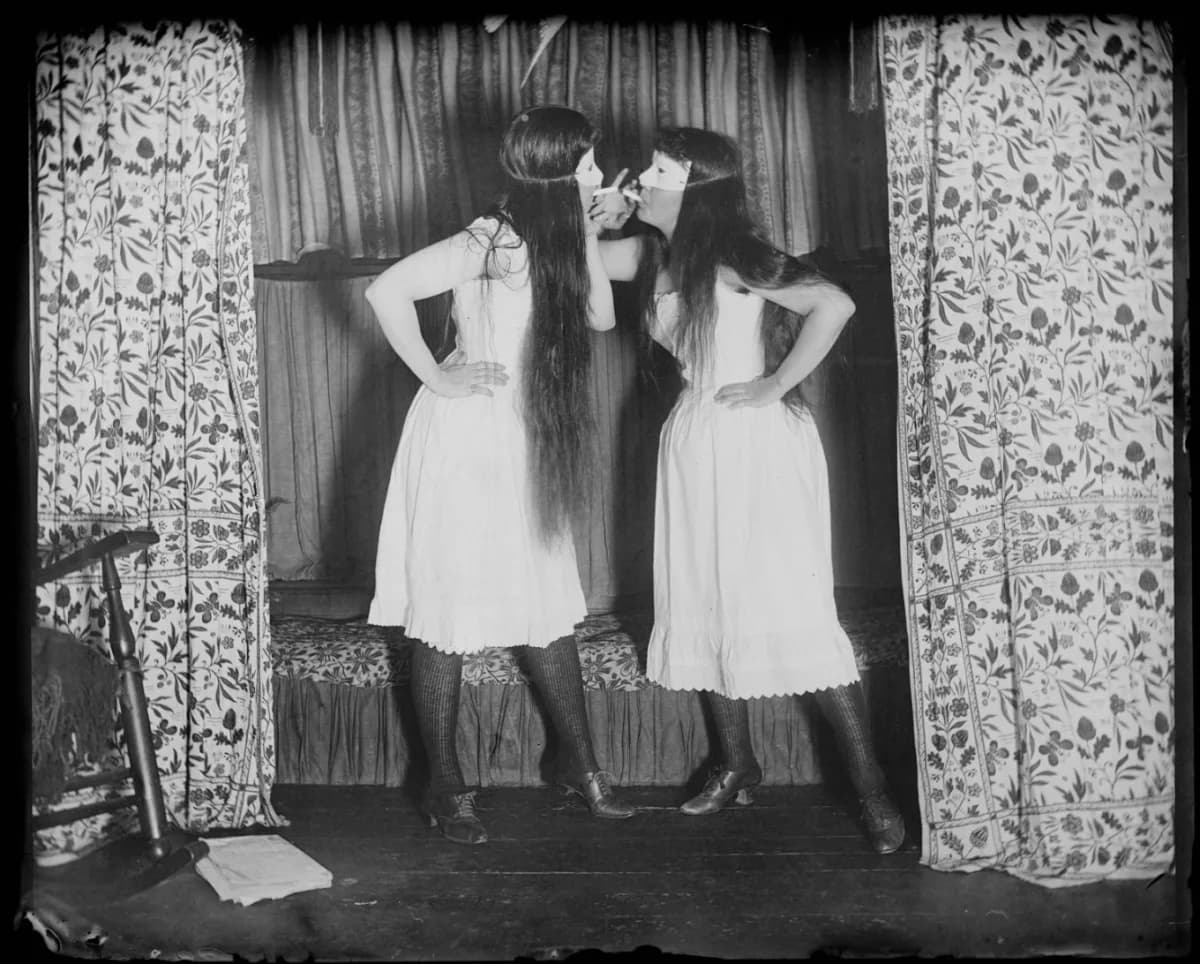 Black and white photograph of two women in white dresses posing with hands on hips against a patterned curtain backdrop, early 20th century.