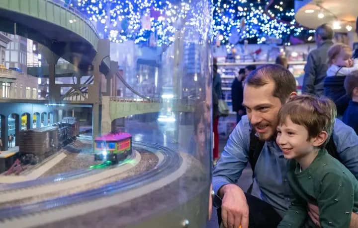 Man and child viewing an illuminated model diorama of a city skyline with blue lighting and architectural details inside a museum.