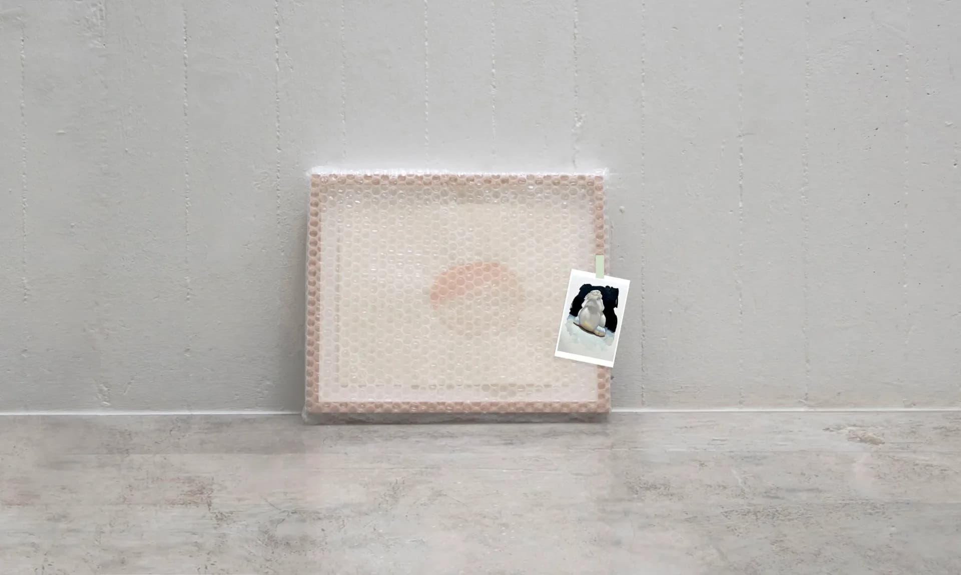 Mixed-media artwork featuring a large cream-colored textured rectangular form with a small photograph or image attached to its right side, displayed against a concrete floor and white wall.