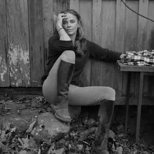 Black and white photograph of a woman in dark jacket sitting outdoors against wooden wall, hand to face.