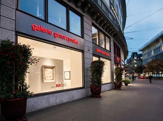 Street-level storefront of Galerie Gmurzynska at dusk, with illuminated interior, red signage, and large display windows on an urban street.