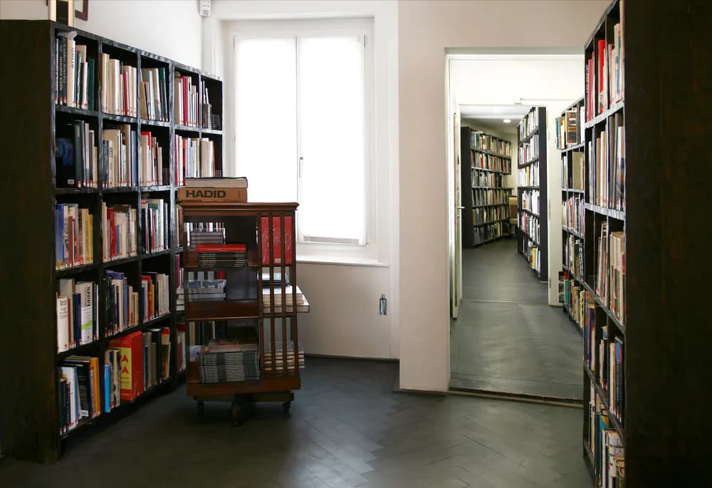 Gallery interior corridor lined with floor-to-ceiling bookshelves on both sides, wooden desk visible on left, neutral lighting.
