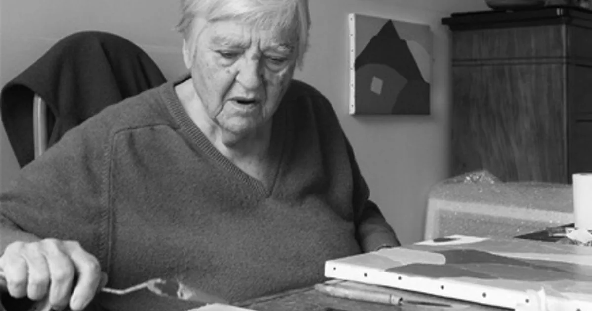 Black and white photograph of an elderly man with white hair leaning over a desk with papers and materials, wearing a dark sweater.