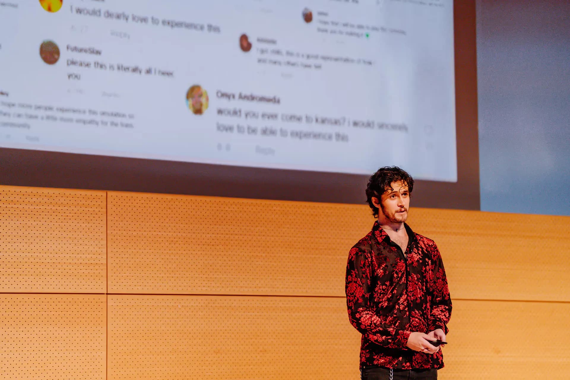 Photograph of a person speaking at a podium in front of a large projection screen displaying social media content in an institutional setting.