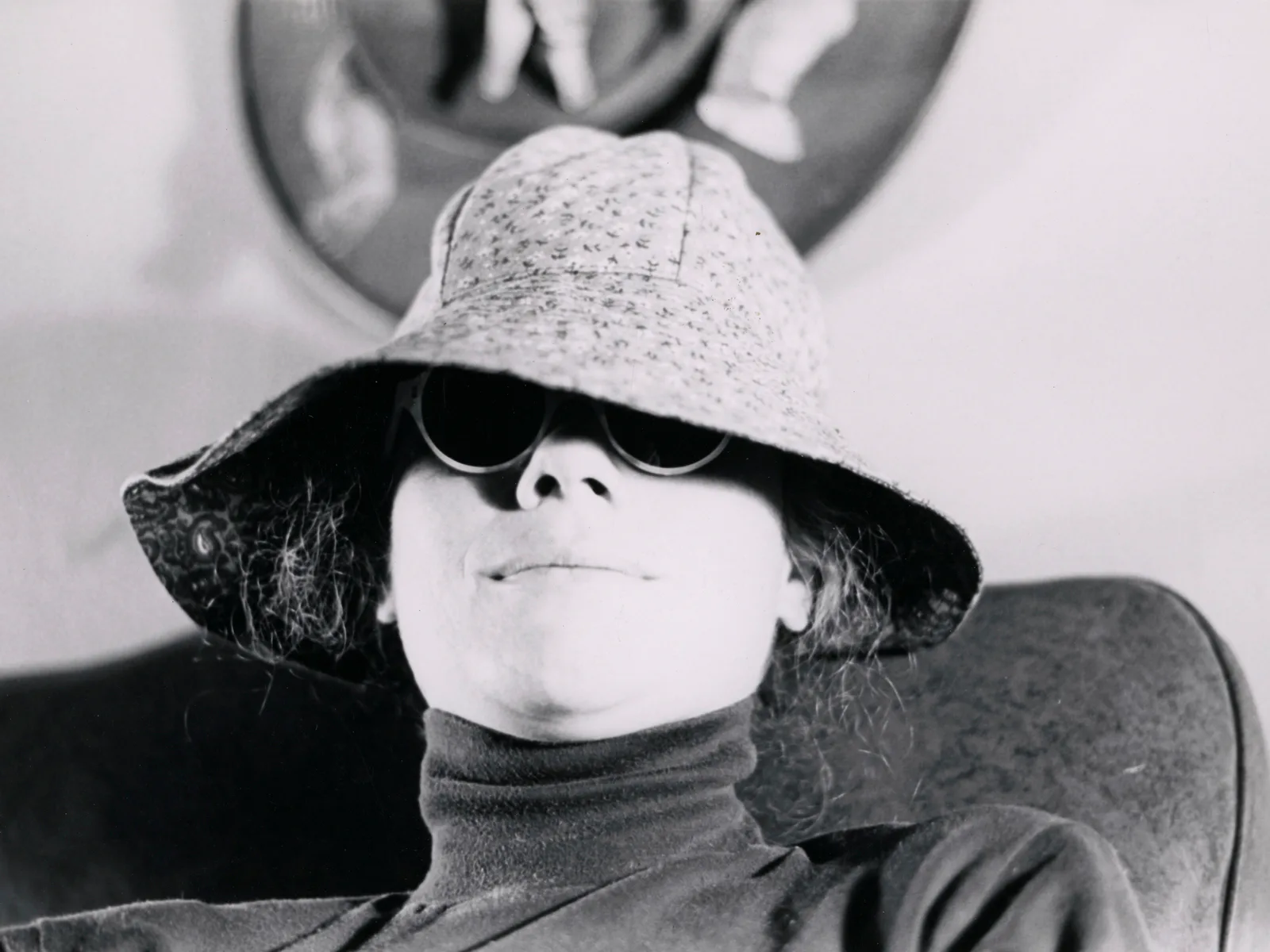 Black and white portrait of a person wearing sunglasses and a wide-brimmed hat with fabric draped behind.