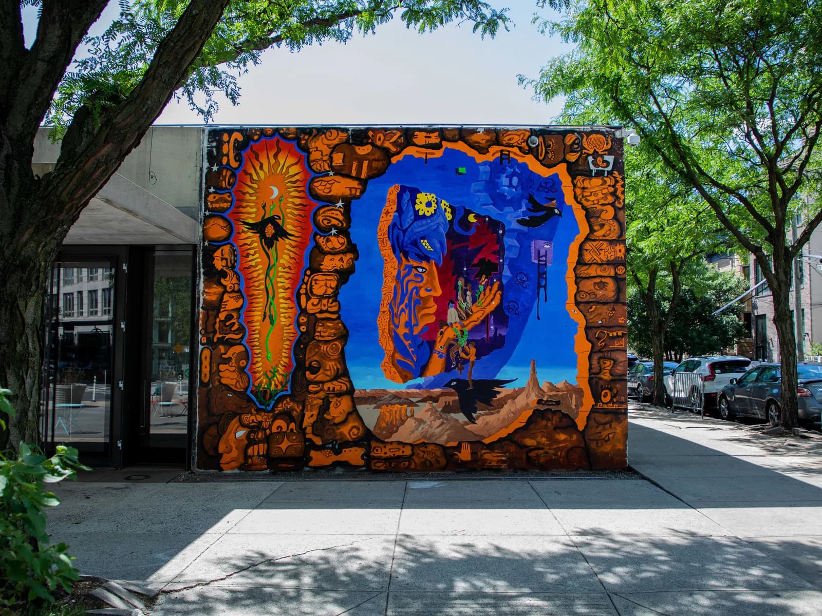 Vibrant mural with orange geometric patterns framing a central blue figure against warm tones, installed on an urban storefront.