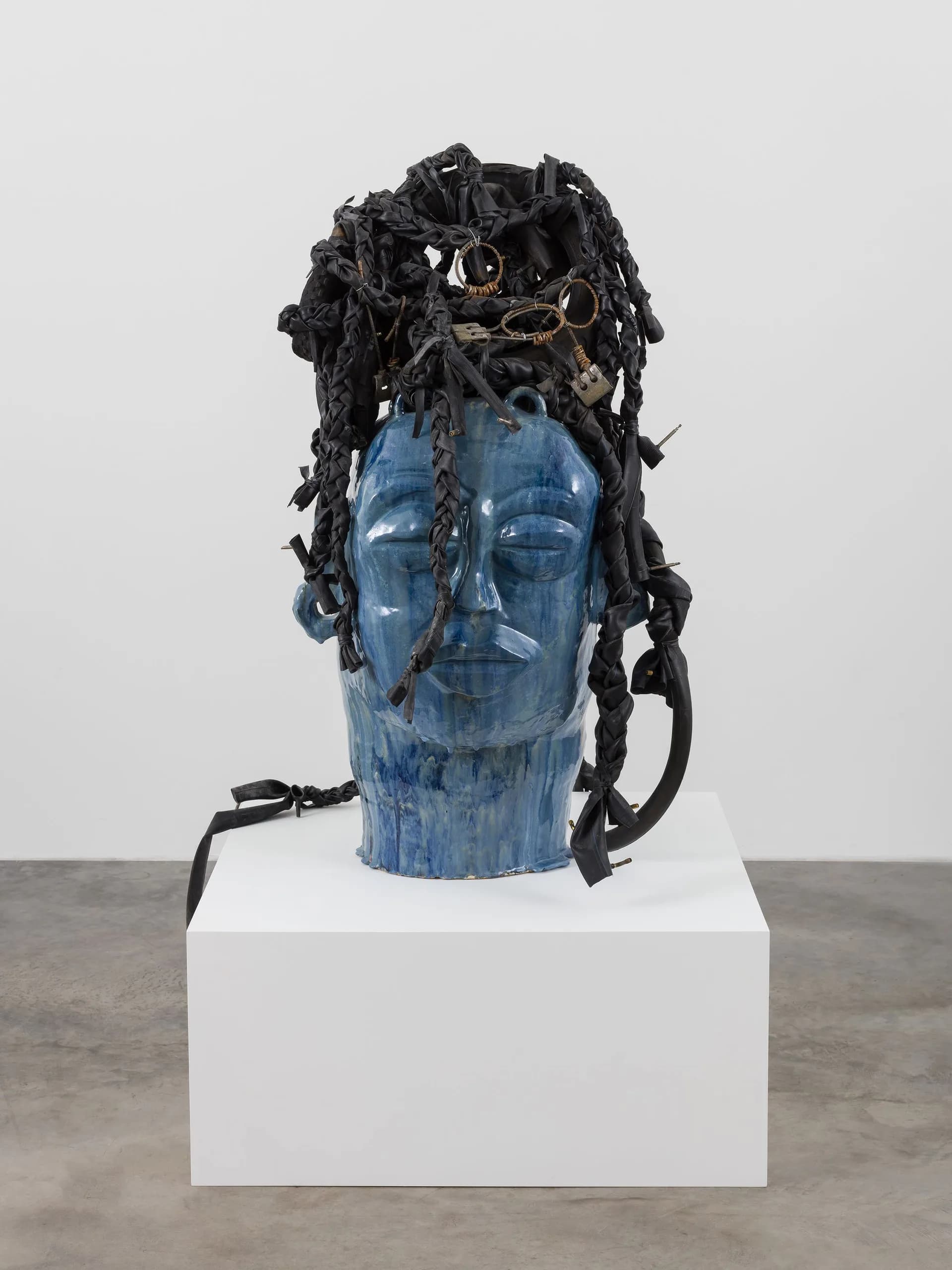 Sculptural head form in blue glazed ceramic mounted on white pedestal, topped with black fabric and wire elements extending upward.