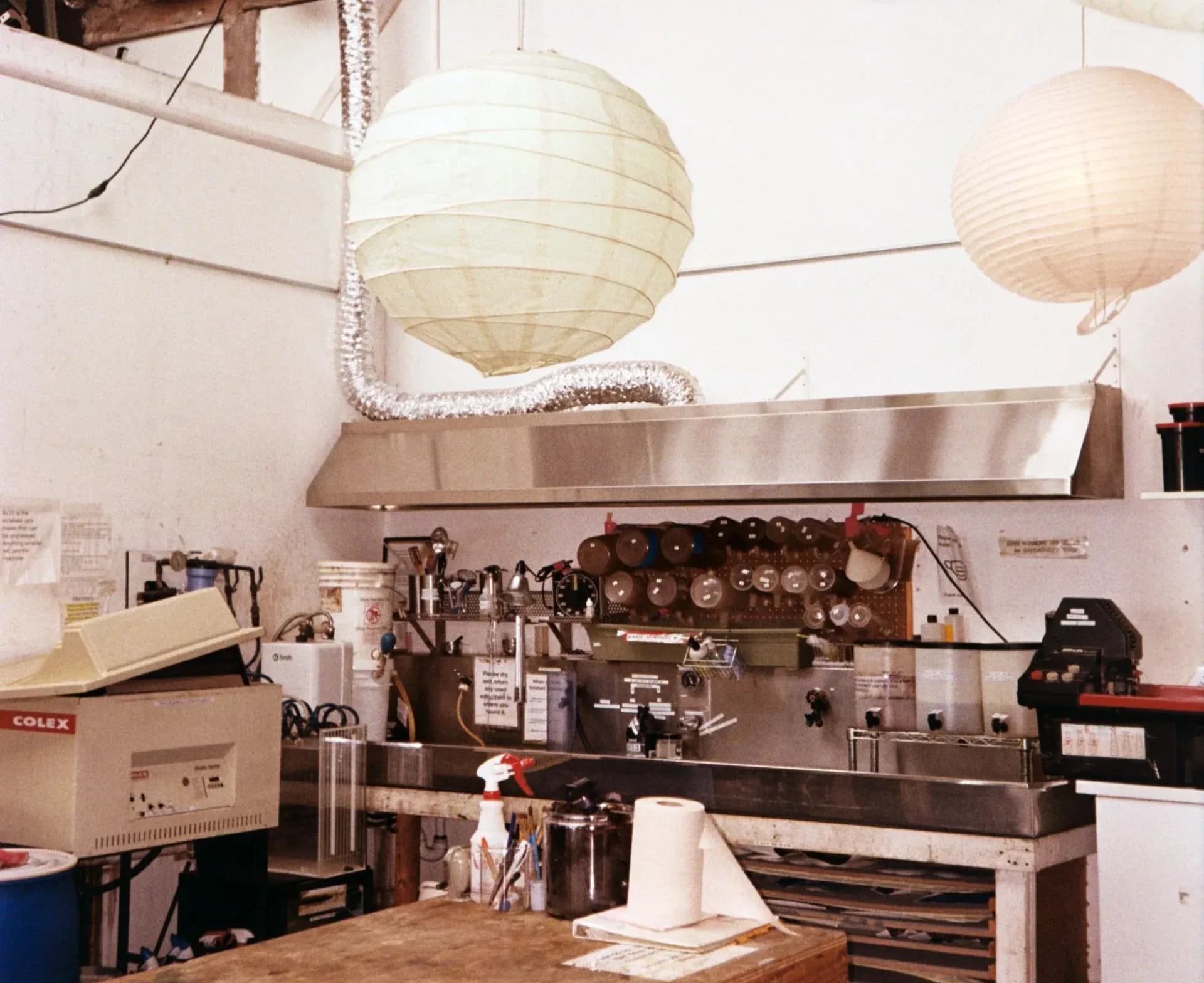 Gowanus Darkroom workspace with stainless steel work surface, hanging paper lanterns, darkroom enlargers, chemical bottles, and processing equipment.