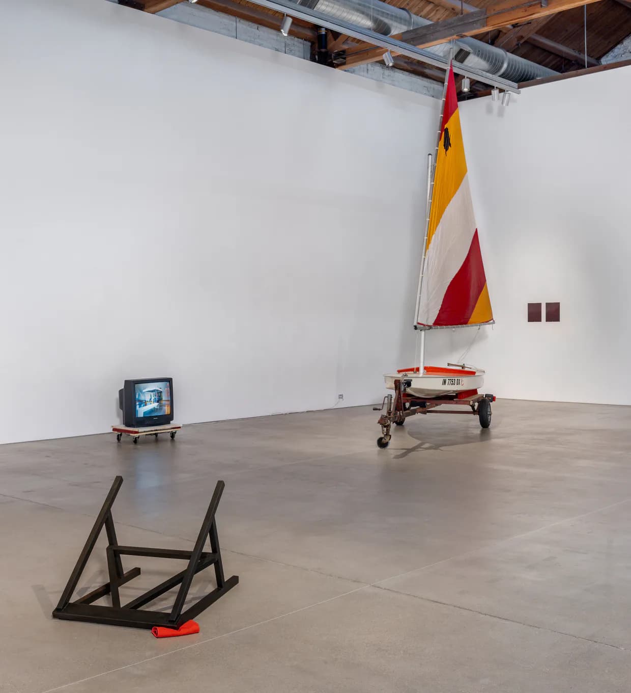 Gallery interior with concrete floor, white walls, sailboat sculpture with red and yellow sail, metal ladder sculpture, and monitor on ground.