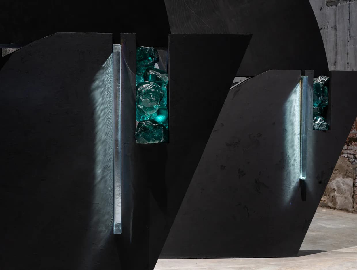 Dark industrial gallery space with angular black walls and vertical display niches containing illuminated turquoise sculptural forms.