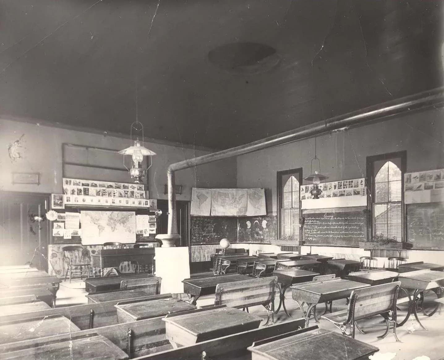 Black and white photograph of a historic schoolroom interior with wooden benches, desks, chalkboards, fireplace, and pendant light fixture.