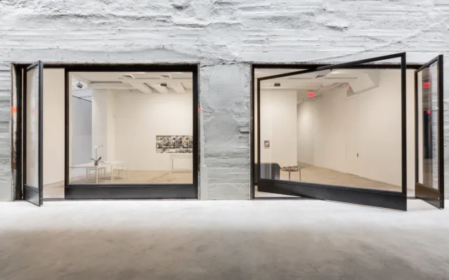 Two minimalist gallery rooms with black-framed glass partitions and cream-colored walls, viewed from a concrete floor.