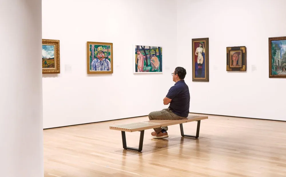 Gallery interior with white walls, wood floor, framed paintings, and a visitor seated on a bench viewing the art