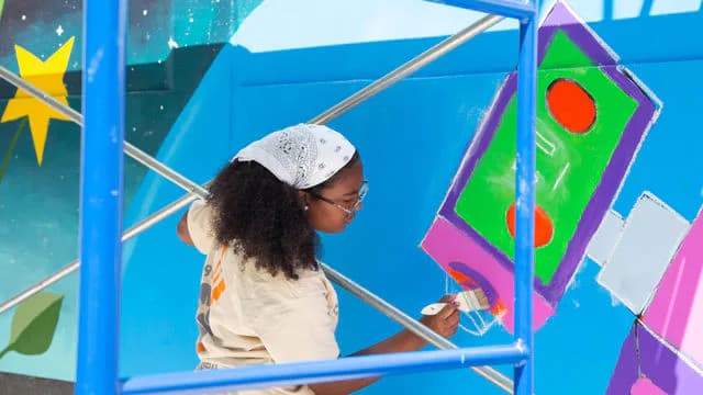 Person painting colorful mural with bright blue, green, pink, and purple geometric shapes and star design on structural framework.