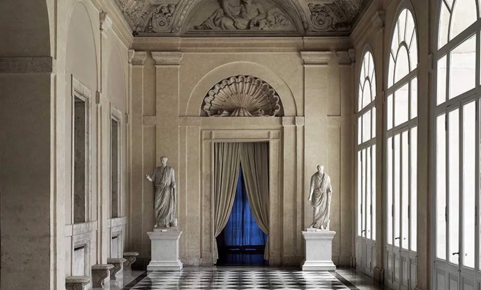 Neoclassical gallery interior with cream walls, white marble floor, sculptural busts on pedestals, and blue-draped artwork at center beneath decorative ceiling.