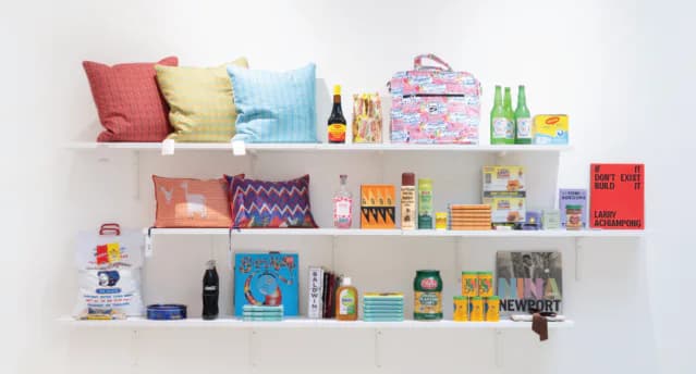 Three white gallery shelves displaying colorful books, household items, cushions, and merchandise in minimalist interior setting.