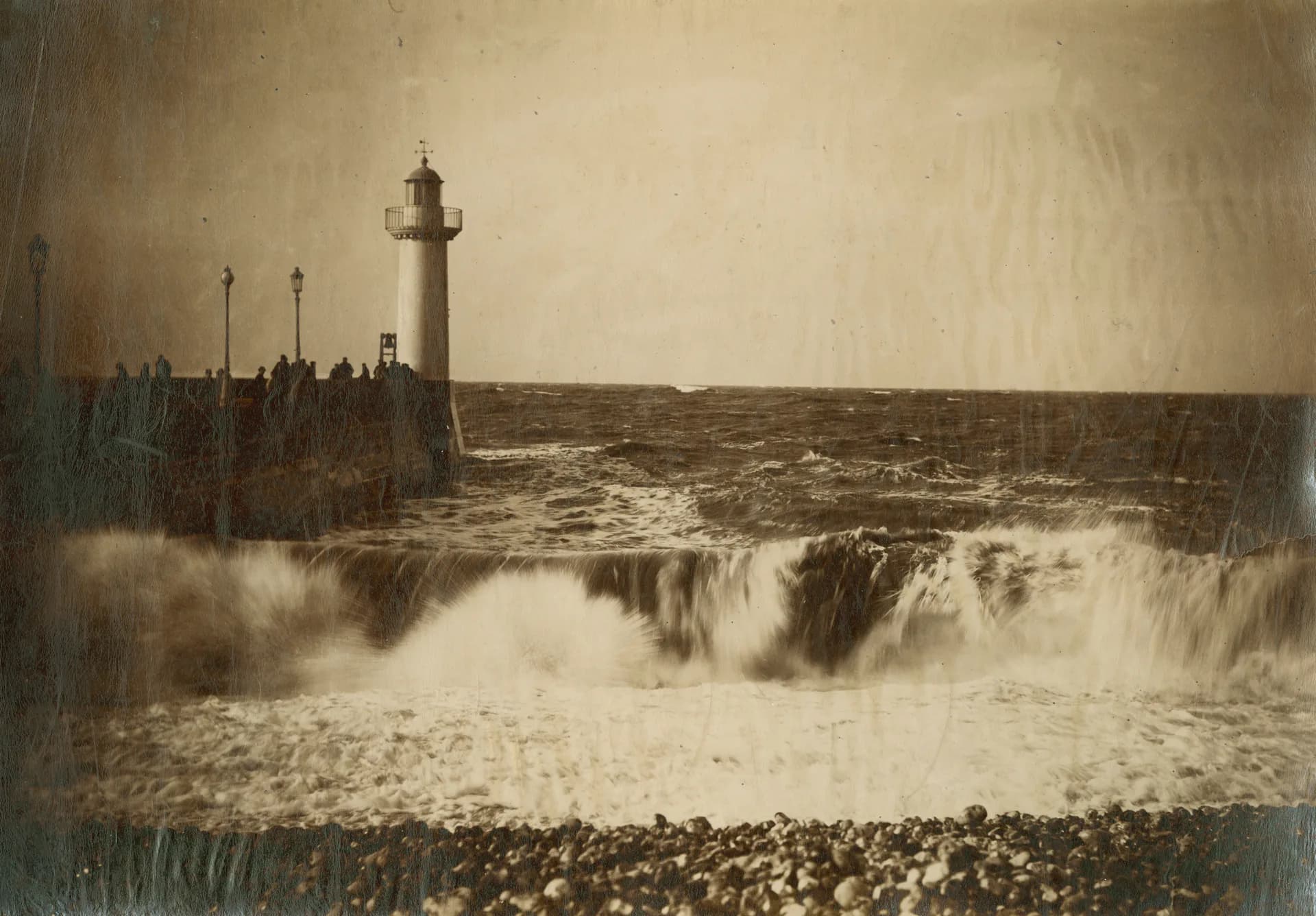 Sepia photograph of a lighthouse on rocky shore with crashing waves and turbulent sky.