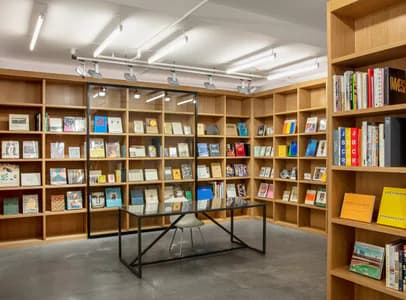Gallery interior with extensive natural wood shelving displaying colorful books and art publications, modern black metal table in center under skylights.