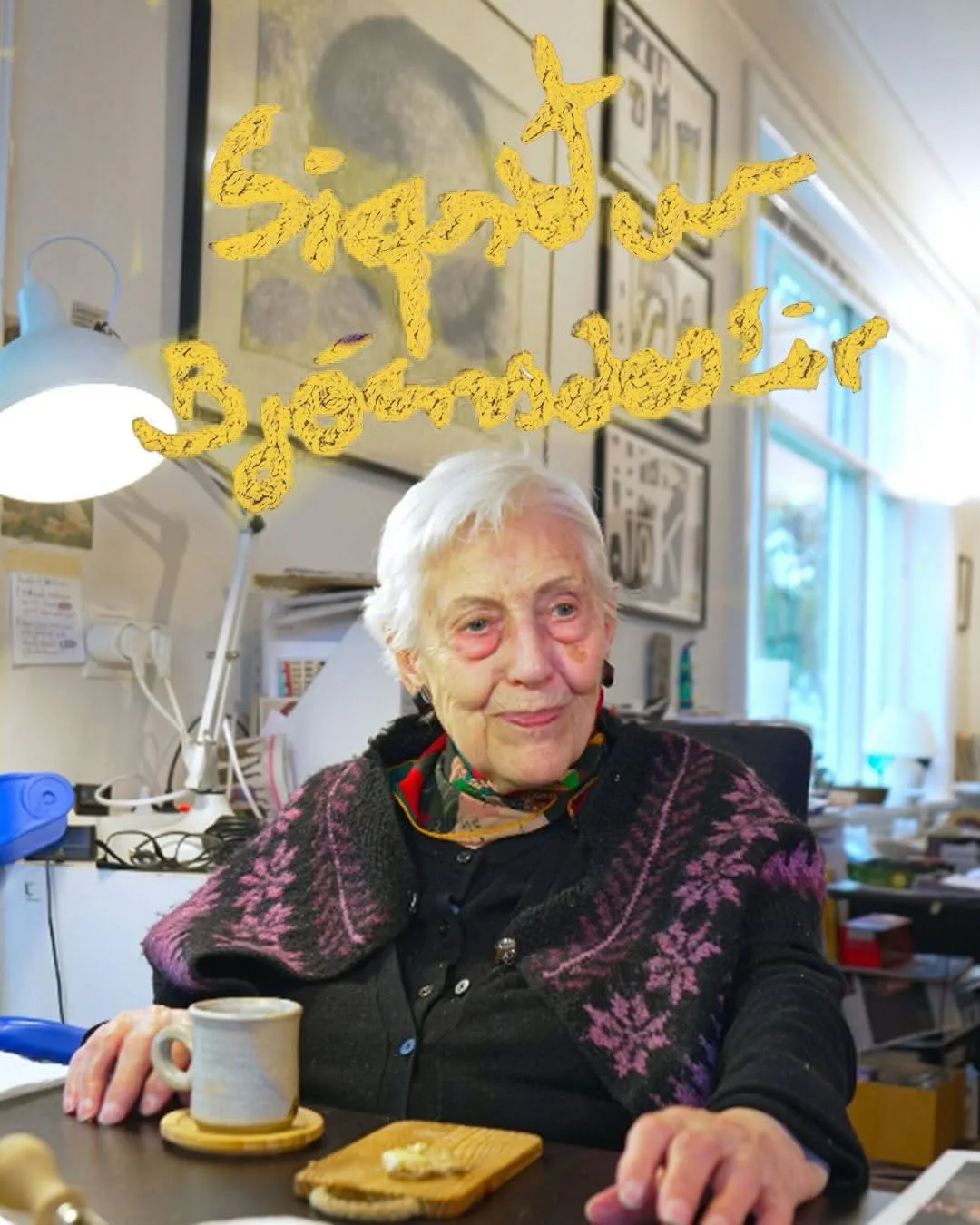 Elderly woman with white hair wearing a patterned purple and black jacket, seated at a desk with a cup and plate in front of her.