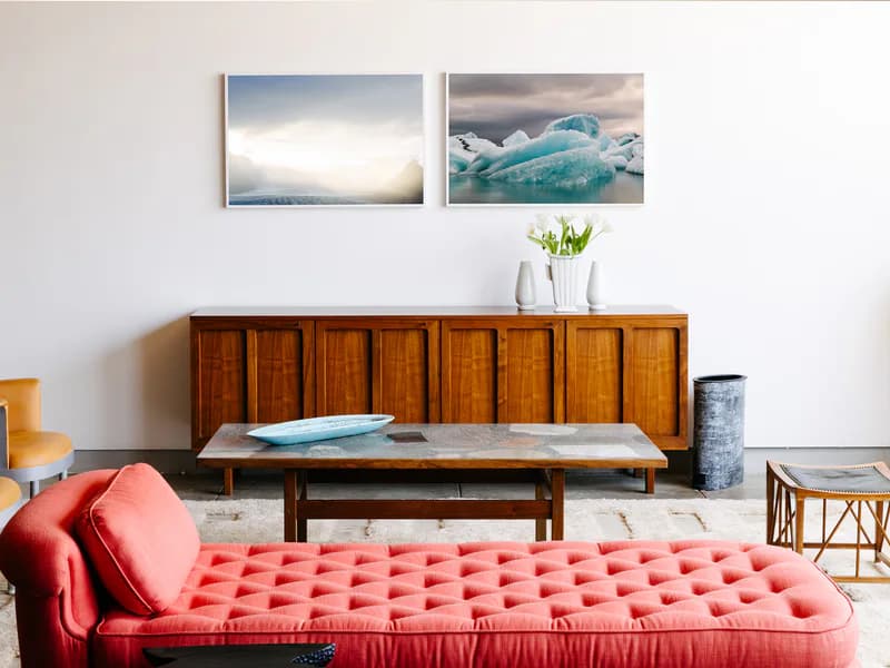 Gallery interior featuring mid-century wooden sideboard, red quilted bench seating, landscape photographs on wall, minimalist furnishings and decor.