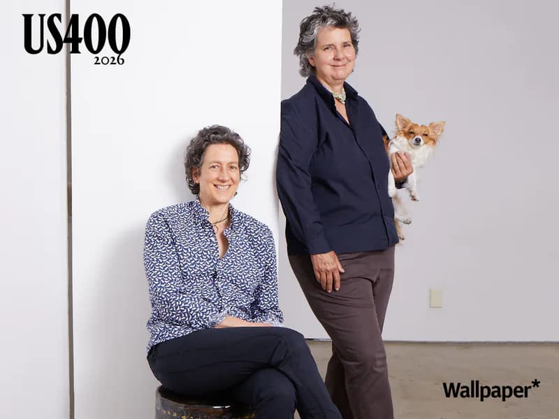 Two women posed indoors, one seated in patterned navy shirt, one standing holding orange cat, with 'US400 2026' and 'Wallpaper*' branding visible.
