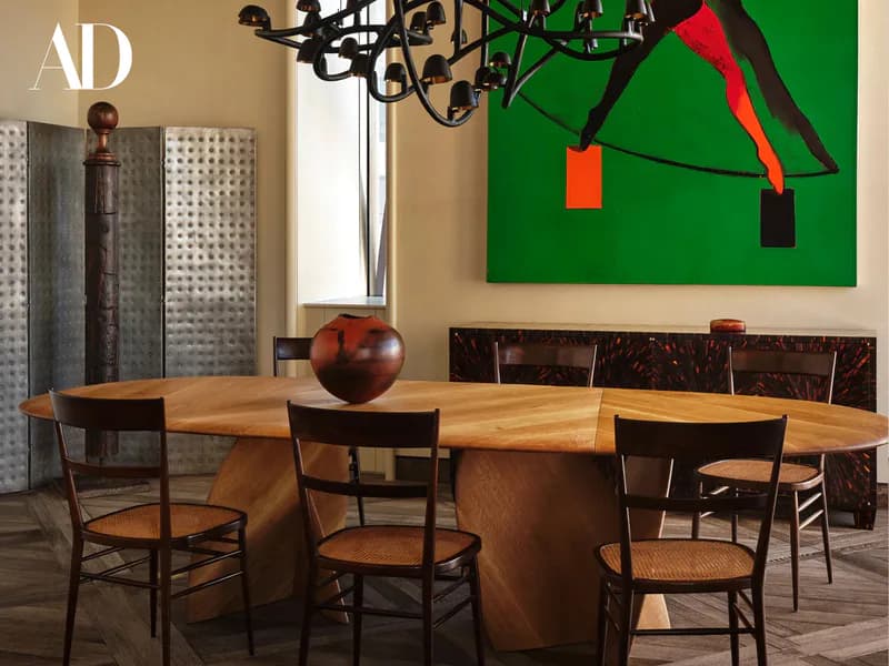Gallery space with wooden dining table and black chairs, green canvas with red sculptural elements, modern chandelier, marble sideboard, 'AD' branding.