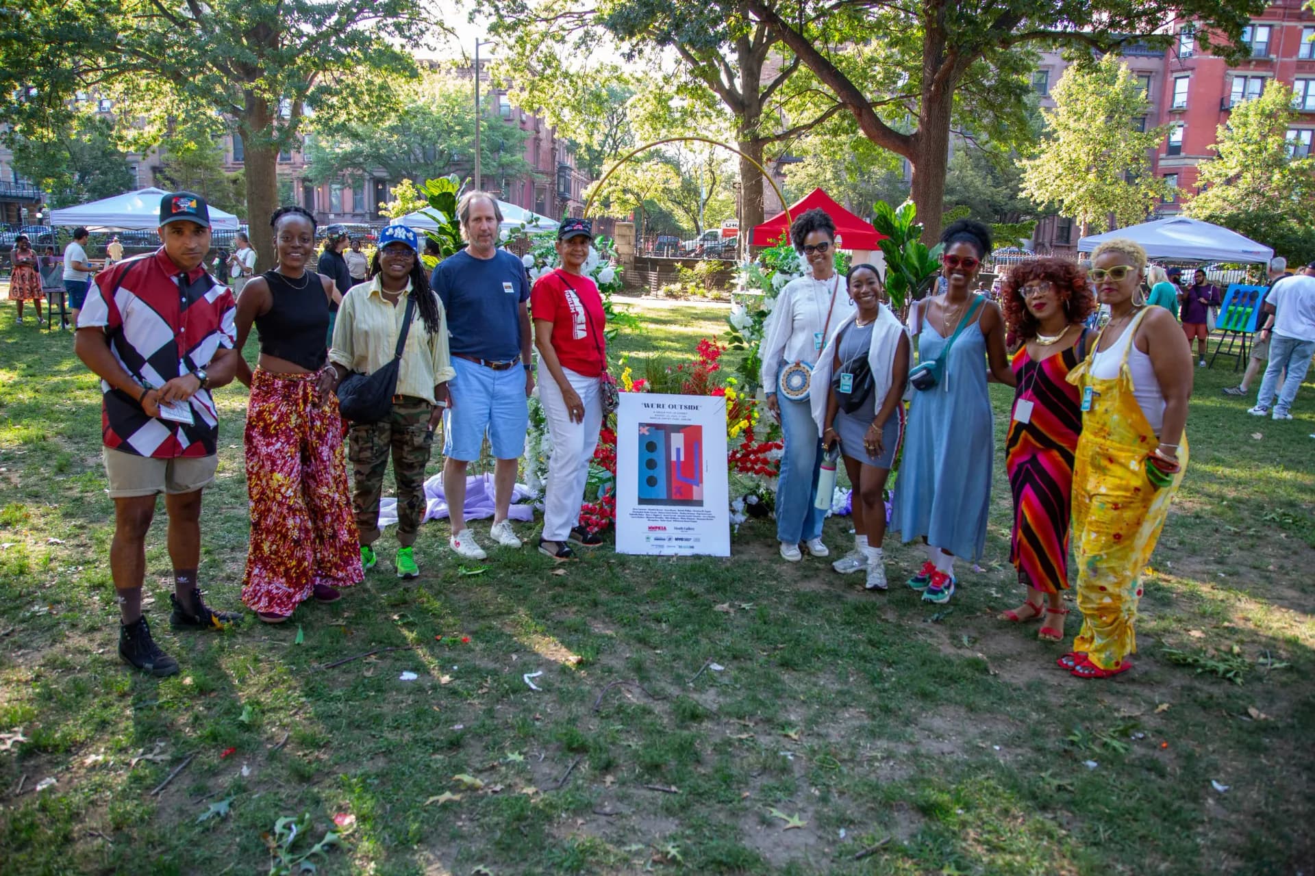 Outdoor community gathering in park with diverse group of people standing around Heath Gallery booth with colorful artwork and tents.
