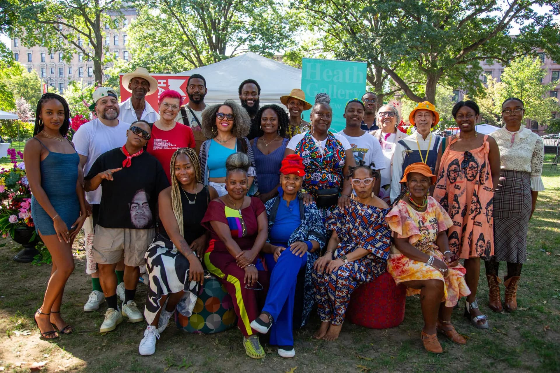 Large group photo of diverse visitors and staff at Heath Gallery outdoor booth in park, wearing colorful clothing and posing together.