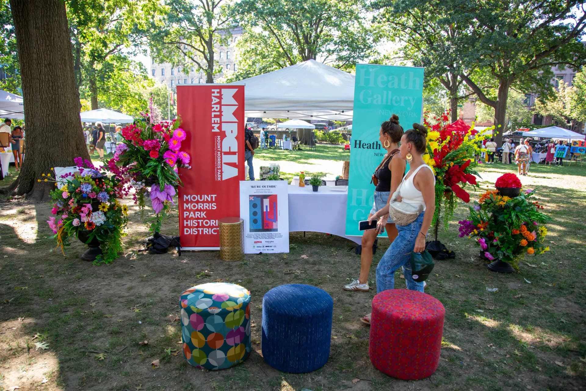 Heath Gallery outdoor booth in park with red and turquoise signage, vibrant floral arrangements, and colorful seating poufs, visitors interacting with staff.