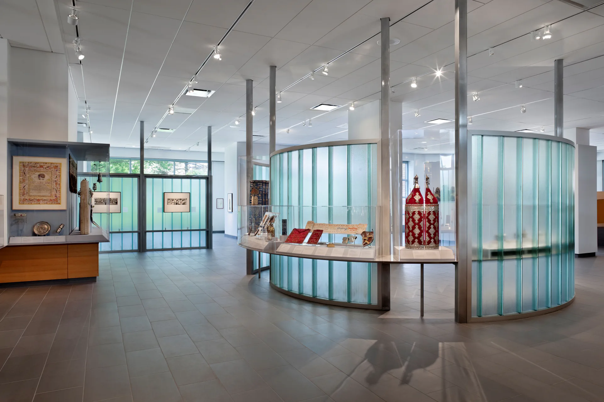 Modern gallery interior with polished floors, structural columns, glass-paneled display cases with turquoise accents, and artwork visible on distant walls.