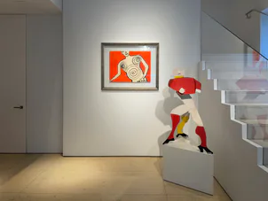 Contemporary gallery space with white partition wall displaying abstract orange artwork, red sculptural figure on pedestal, and staircase visible.