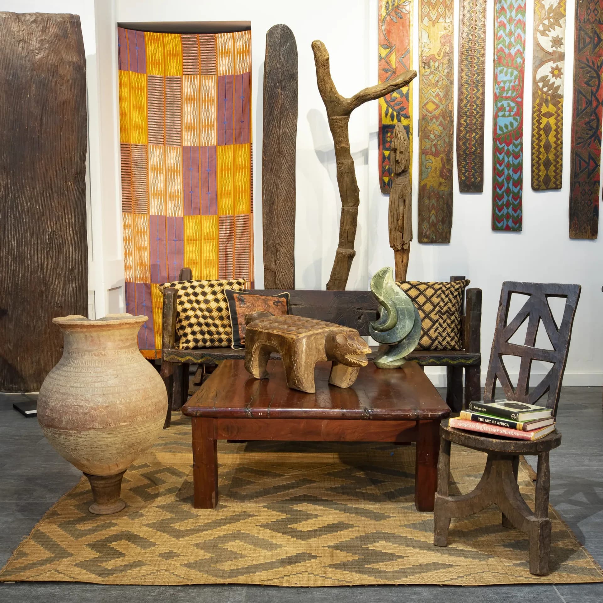 Hemingway Gallery interior displaying African-inspired furniture and textiles: wooden bench with patterned cushions, carved wooden sculptures, woven baskets, and decorative wall-mounted fabric panels in earth tones.