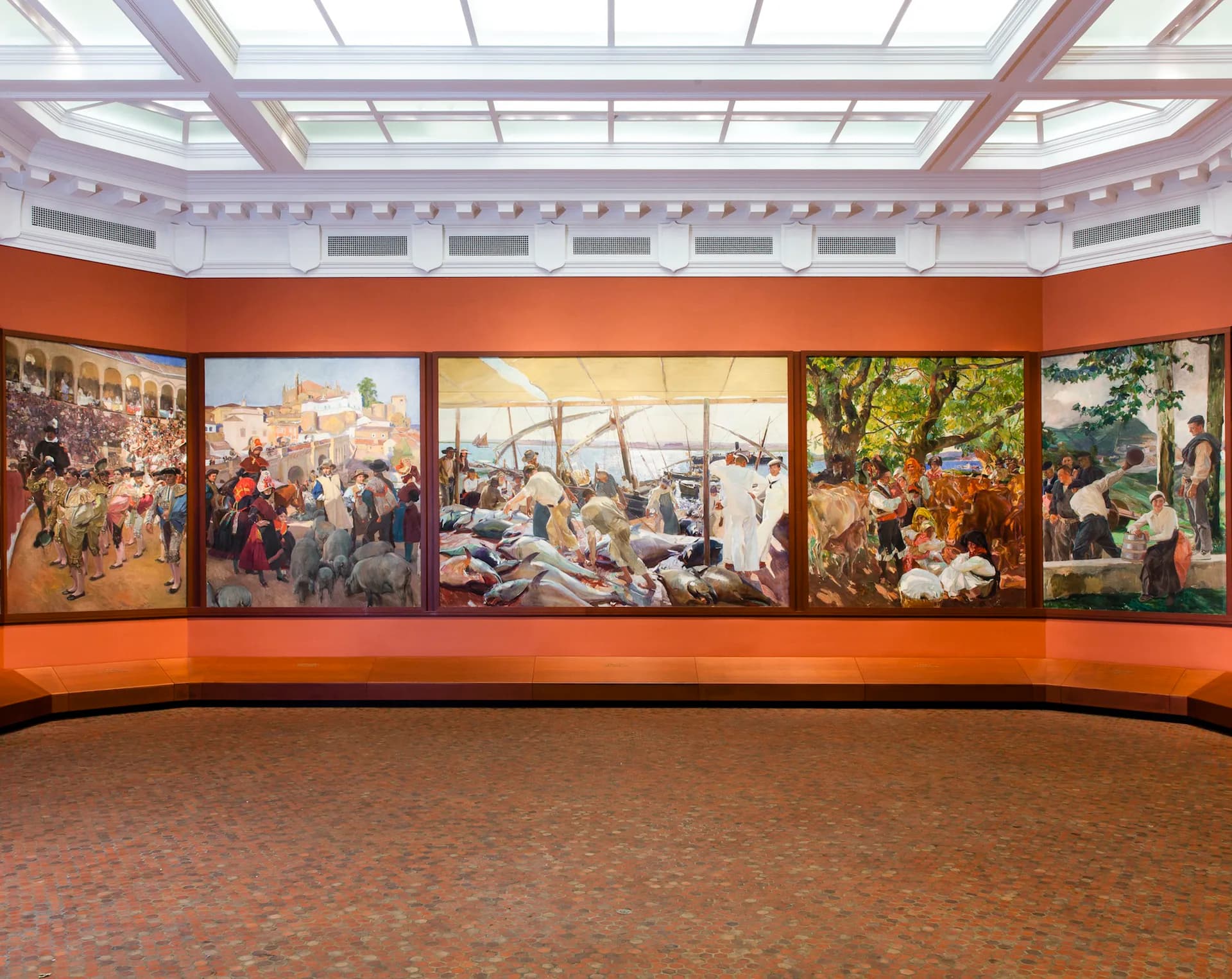 Museum gallery with curved orange walls displaying five large historical paintings depicting scenes of daily life, with skylights above and terracotta flooring.