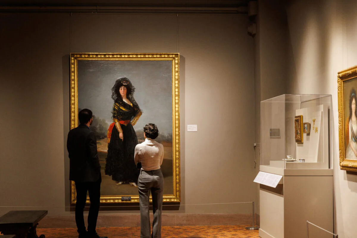 Gallery interior with visitors viewing a large framed portrait painting of a woman in black clothing against a gray wall with gold frame.